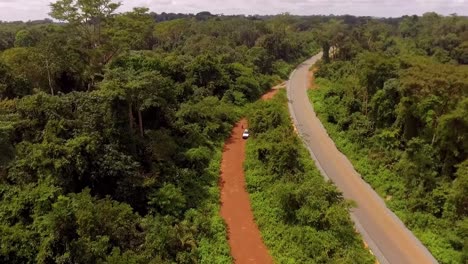 Aerial-drone-view-away-from-a-car,-revealing-a-rainforest-road-and-the-Jungle,-on-a-sunny-day,-in-Nanga-Eboko,-Haute-Sanaga,-Southern-Cameroon