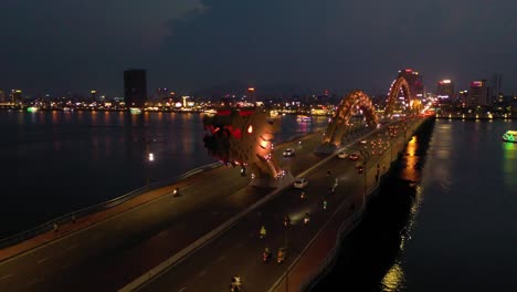 Drone-spinning-around-head-of-Dragon-Bridge-Cau-Rong,-traffic-and-city-skyline-at-night-in-Danang,-Vietnam