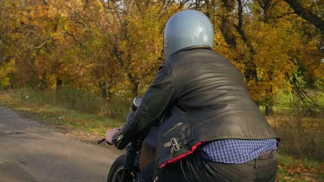 Rückansicht-Eines-Mannes-Mit-Grauem-Helm,-Lederjacke-Und-Kariertem-Hemd,-Der-An-Einem-Sonnigen-Herbsttag-Auf-Einer-Asphaltstraße-Motorrad-Fährt