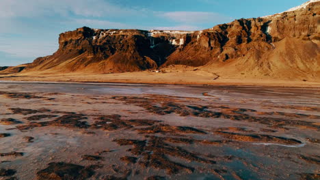 aerial view flying across icy reynisfjara black sand beach towards golden sunlit icelandic mountains