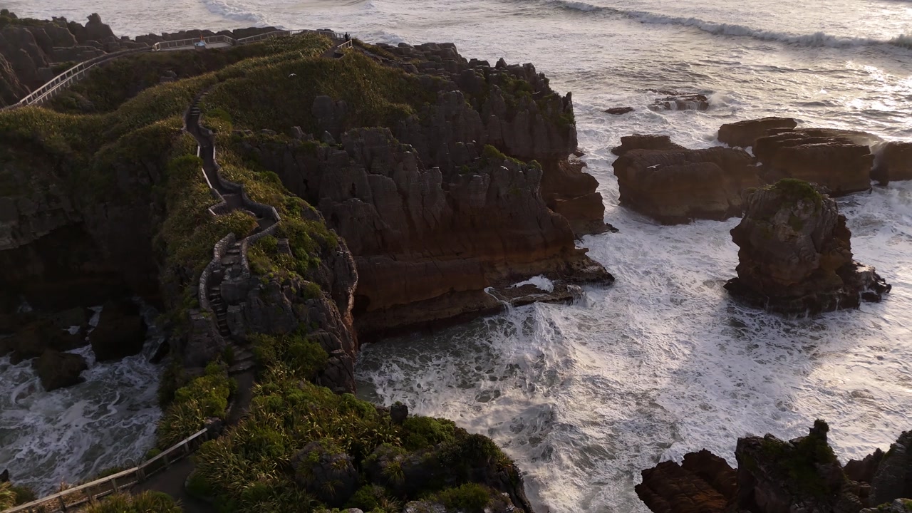Aerial view of Pancake Rocks in Punakaiki on West Coast of New Zealand’s South Island, with waves crashing against rugged cliffs and seabirds flying in warm sunset light.