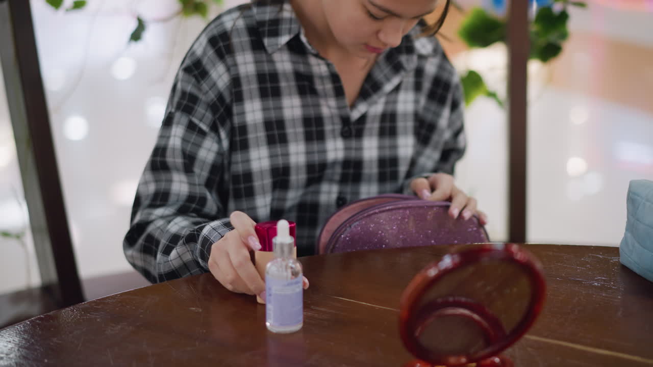 카레드 셔츠를 입은 젊은 여성이 나무 테이블, 나무 거울, 부드러운 배경 조명 위에 반이는 보라색 화장품 가방에서 화장품을 가져옵니다.