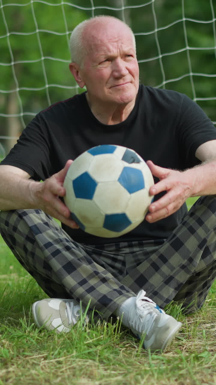 An elderly man sits cross-legged near a goal post, gently toggling a soccer ball in his hands while looking around, with a blue pole in the background and trees