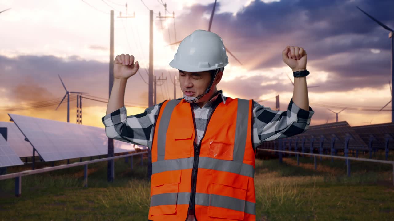 Asian Male Engineer With Safety Helmet Screaming Goal And Dancing Celebrating With Solar Panel and Wind Turbines