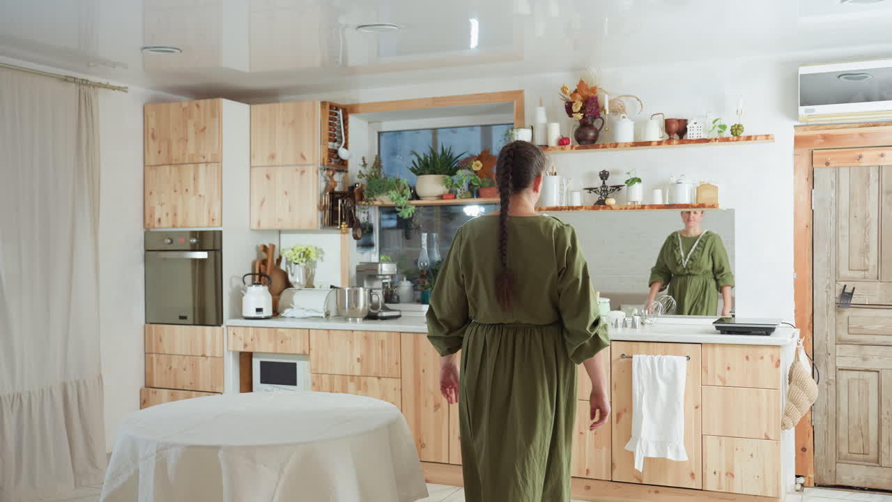 Woman in green dress with braided hair walks into kitchen decorated with wood cabinets, indoor plants, utensils, oven, and mirror reflecting her front view as she prepares to begin household activity