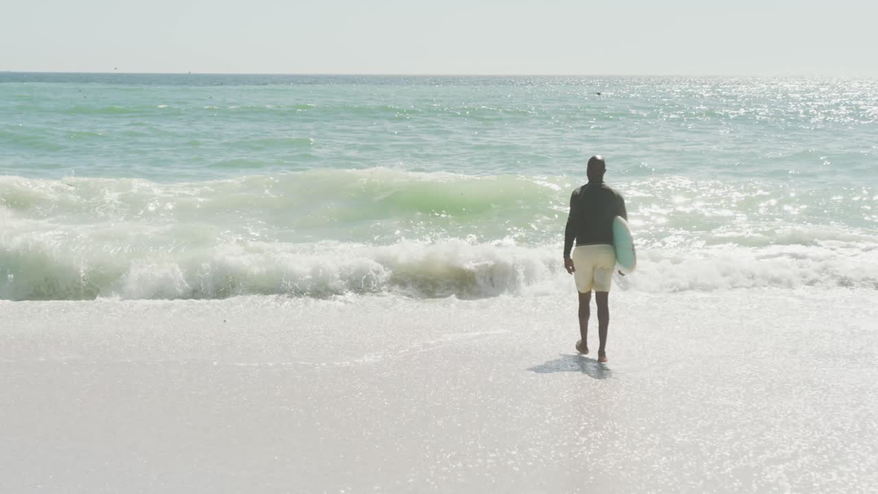 시니어 아프리카계 미국인 남자가 서핑 보드와 함께  ⁇ 은 해변을  ⁇ 고 있습니다.