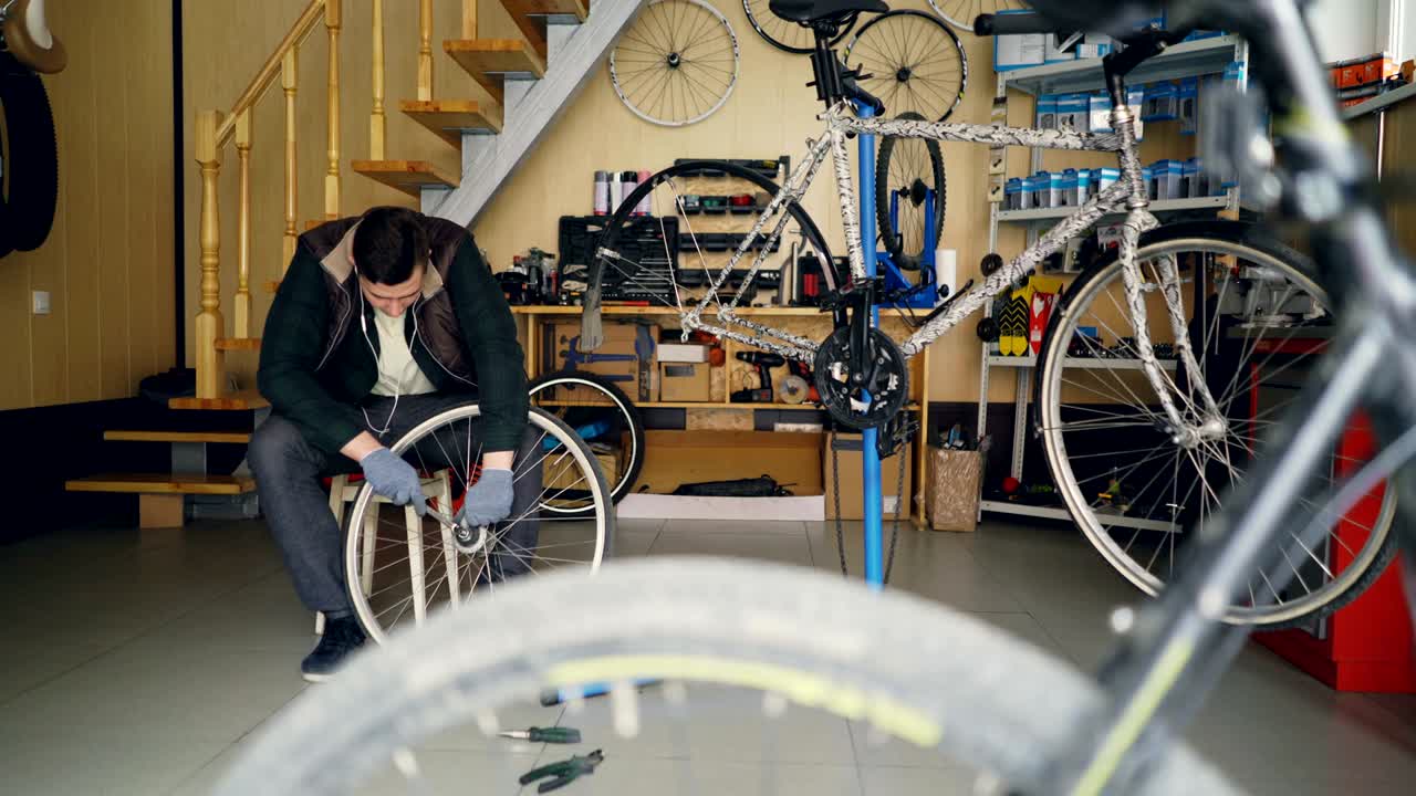 Handsome mechanic is fixing back wheel with wrench while working in modern workplace sitting on wooden stool. Cycles, spare parts, tools and equipment are visible.