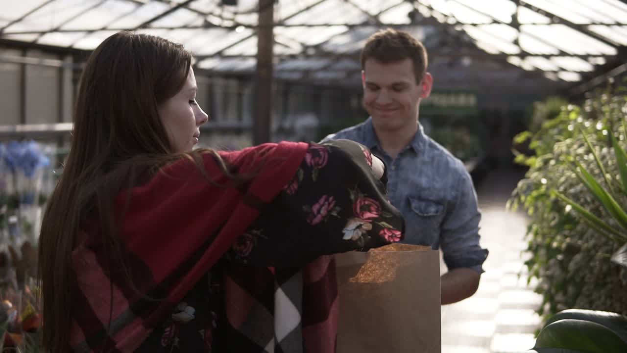 판매 여성이 온실에서 고객에게 유기농 음식을 제공하고 있는 희귀한 모습. 여성은 녹색, 과일 및 채소를 갈색 종이 봉투에 포장하고 있습니다. 즐거운 남성 고객이 수확 시장에 왔습니다. 사람들과 건강한 생활 방식 개념