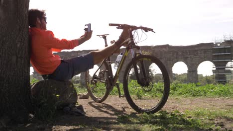 ciclista con bicicleta sentado en una roca bajo un árbol tomando selfies frente al antiguo acueducto romano. joven atractivo hombre atlético con ropa deportiva naranja y mochila en el parco degli acquedotti en roma