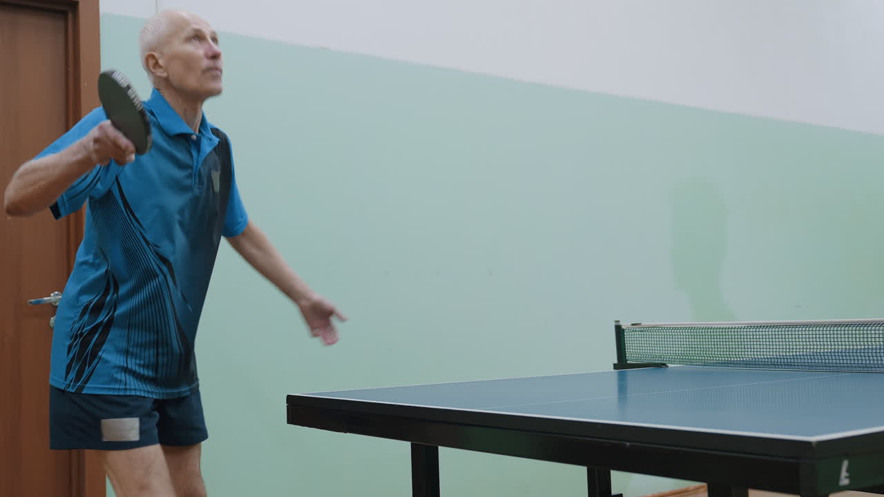 Elderly man in blue sportswear serves tennis ball on indoor table while opponent shadow appears on green wall, highlighting intensity of competitive match and atmosphere of focused gameplay