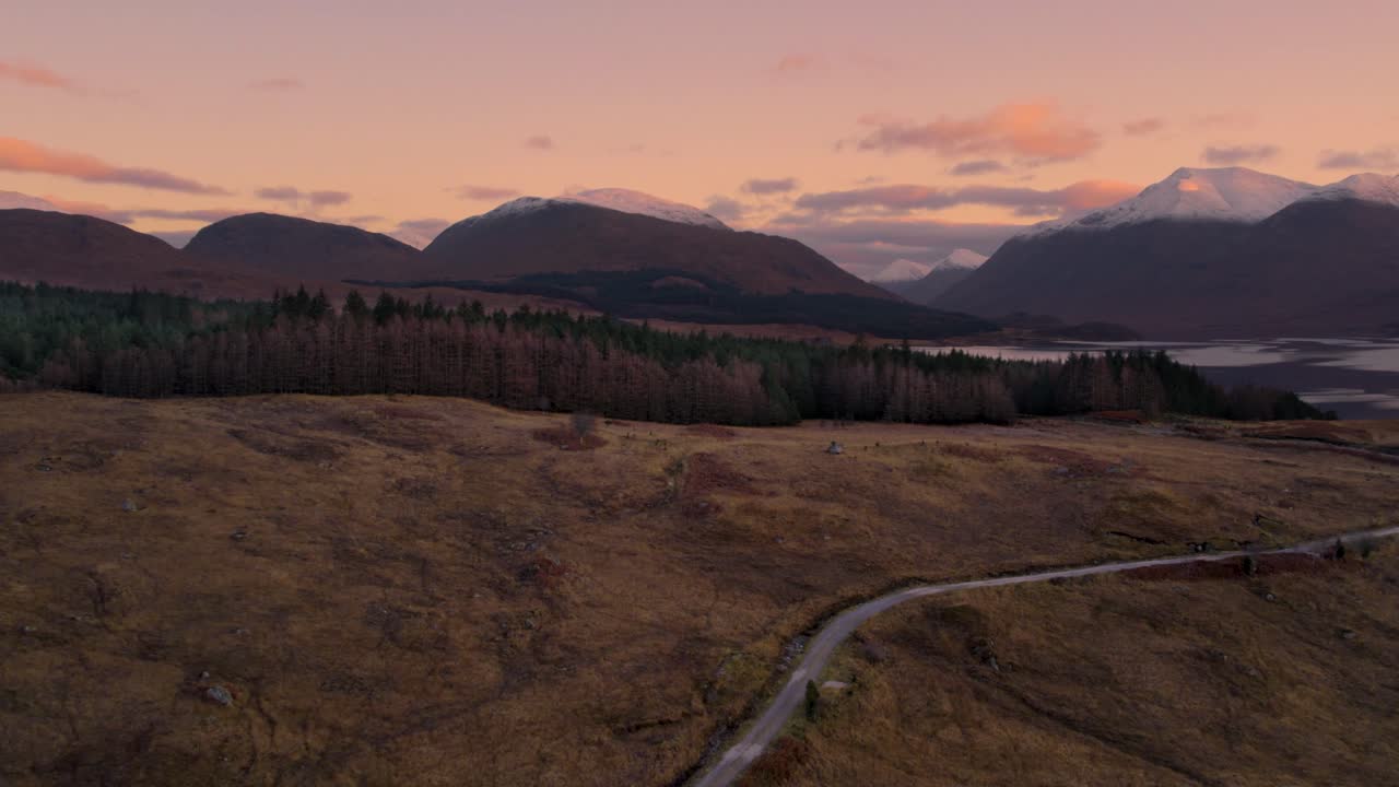 Aerial drone footage flying away from Glen Etive and Loch Etive in Scotland during an orange and pink sunrise of pastel colours, with snow-capped mountains and a forest in the background