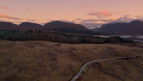 aerial drone footage flying away from glen etive and loch etive in scotland during an orange and pink sunrise of pastel colours, with snow-capped mountains and a forest in the background