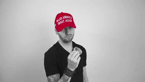 grayscale footage of a man with tattoos, short hair and a beard wearing a red colored maga make america great again hat and lifting up reflective and glossy bitcoin cryptocurrency coins, man is happy.
