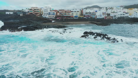 Vista-Aérea-En-órbita-Sobre-La-Playa-Y-Casas-Ubicadas-En-La-Costa-De-El-Puertillo-En-El-Norte-De-La-Isla-De-Gran-Canaria-Y-Con-Las-Olas-Golpeando-Las-Rocas.