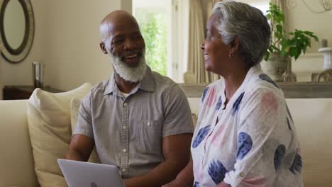 Afroamerikanisches-älteres-Paar,-Das-Einander-Ansieht-Und-Lächelt,-Während-Es-Zu-Hause-Gemeinsam-Einen-Laptop-Benutzt