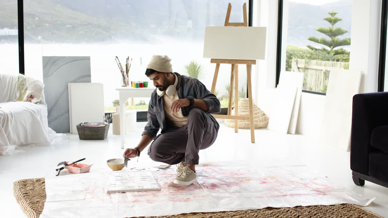 Indian man picking paintbrush from bowl and painting block pattern on floor canvas in studio