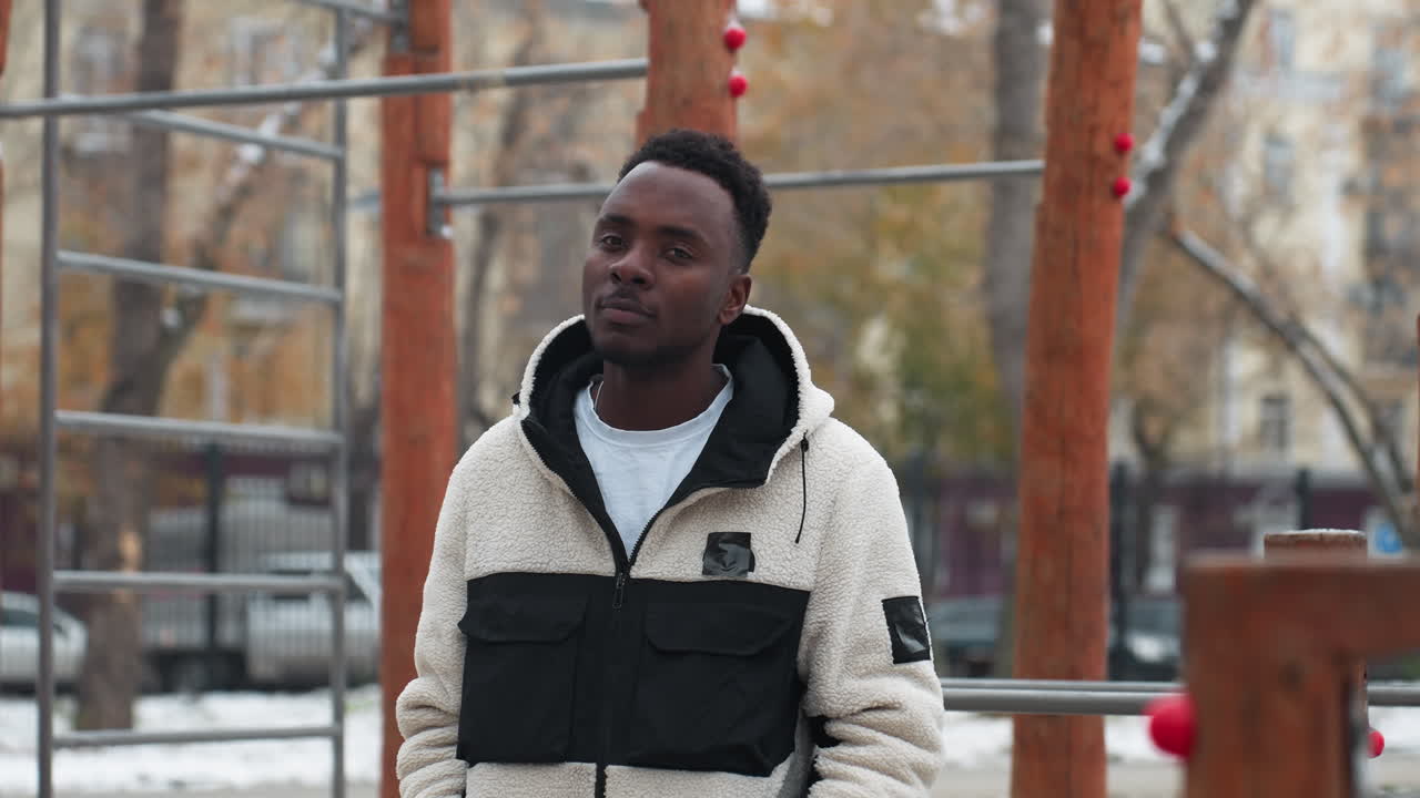 Un uomo africano allegro con una calda giacca invernale ride mentre il fiato freddo è visibile nell'aria gelida. Terreno innevato e alberi spogli creano un'atmosfera invernale suggestiva in un ambiente urbano all'aperto.