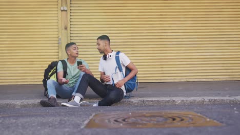 dos felices amigos varones de raza mixta sentados, usando un teléfono inteligente en la calle