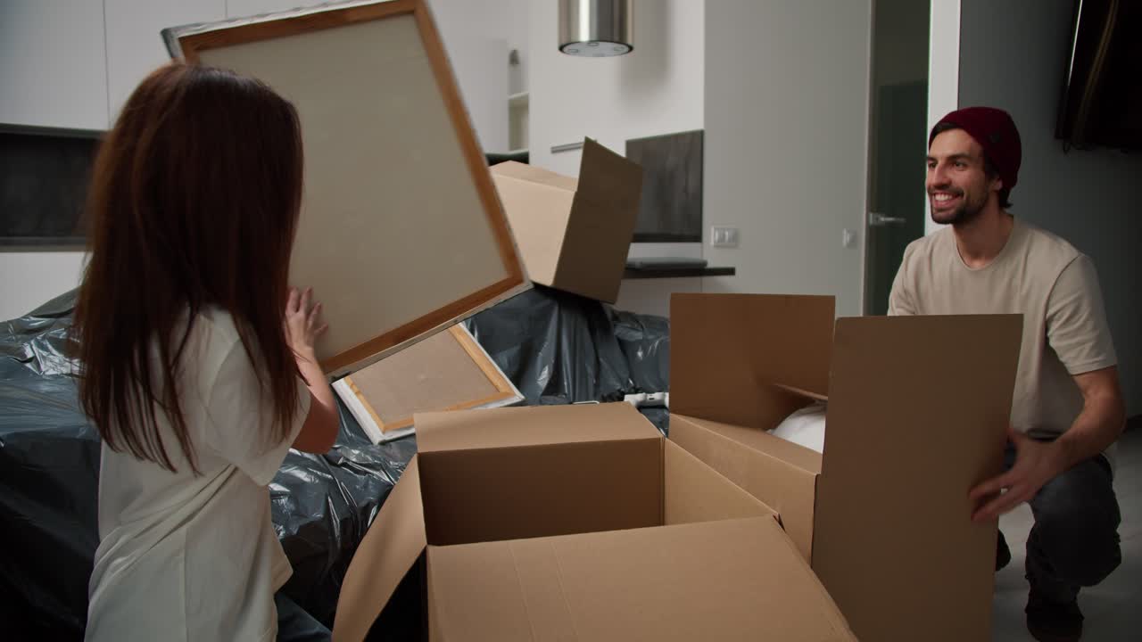 A happy brunette man with stubble in a beige T-shirt brings things in a special box for his brunette girlfriend in a white T-shirt in a new apartment they are sorting things out of the boxes and settling in after moving