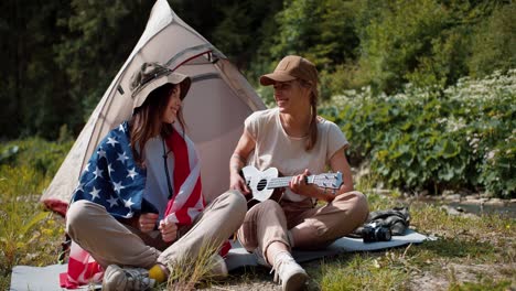 A-brunette-girl-is-wrapped-in-the-flag-of-the-United-States-of-America-and-a-girl-in-camping-clothes-plays-the-guitar,-they-are-sitting-near-the-tent-against-the-backdrop-of-a-green-forest