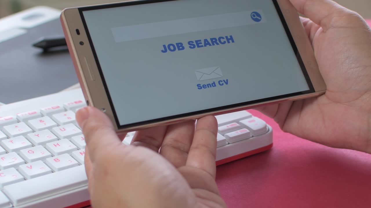 Online job search, woman's hands holding a mobile in which an employment website appears. Work desk with keyboard in the background.