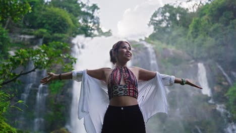 woman smiling and posing confidently in front of a large waterfall, wearing a patterned top and black shorts