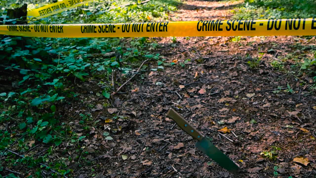 Shiny kitchen knife stabbed in a hiking trail in front of a yellow colored Crime Scene police tape ribbon during a sunny day at a rural countryside forest with brown leaves on the ground. Autumn day