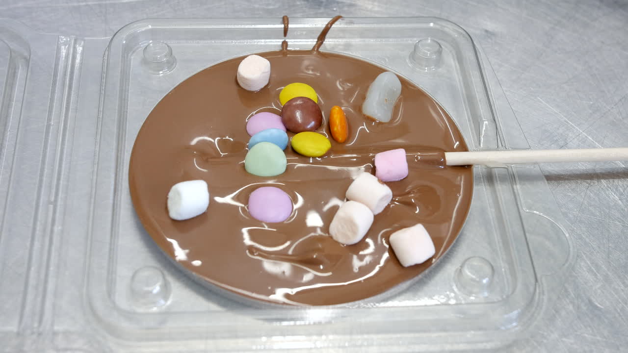 Un lecca-lecca rotondo al cioccolato viene decorato con caramelle colorate e mini marshmallow. Il cioccolato fuso si trova in uno stampo trasparente con un bastoncino di legno posizionato su un lato.