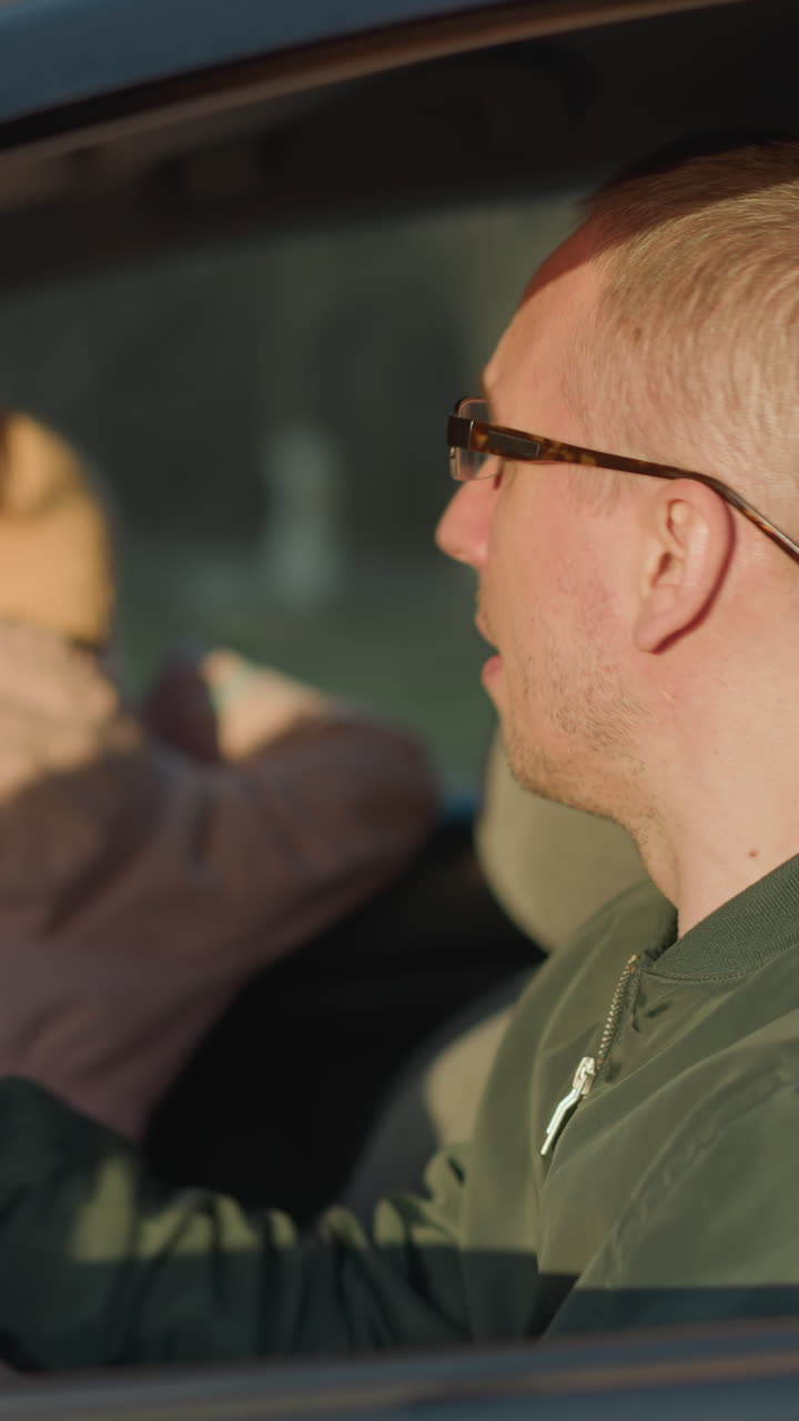 un hombre con una chaqueta verde se sienta en un coche, ajustando sus gafas mientras una chica joven con una jaqueta rosa y un sombrero de punto mira por la ventana