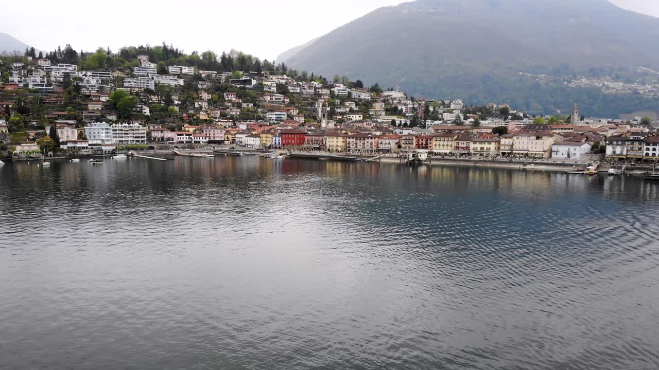 sobrevuelo aéreo sobre la orilla del lago maggiore hacia el paseo junto al lago de ascona, suiza, con vistas a los tejados de la ciudad, la torre de la iglesia y las montañas al fondo
