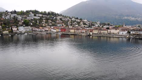 Aerial-flyover-over-the-shore-of-Lago-Maggiore-towards-the-lakeside-promenade-of-Ascona,-Switzerland-with-a-view-of-the-city-rooftops,-church-tower-and-mountains-in-the-background