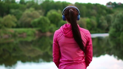 a girl in a pink jacket and blue headphones stands with her back and looks at the river early in the morning after a run