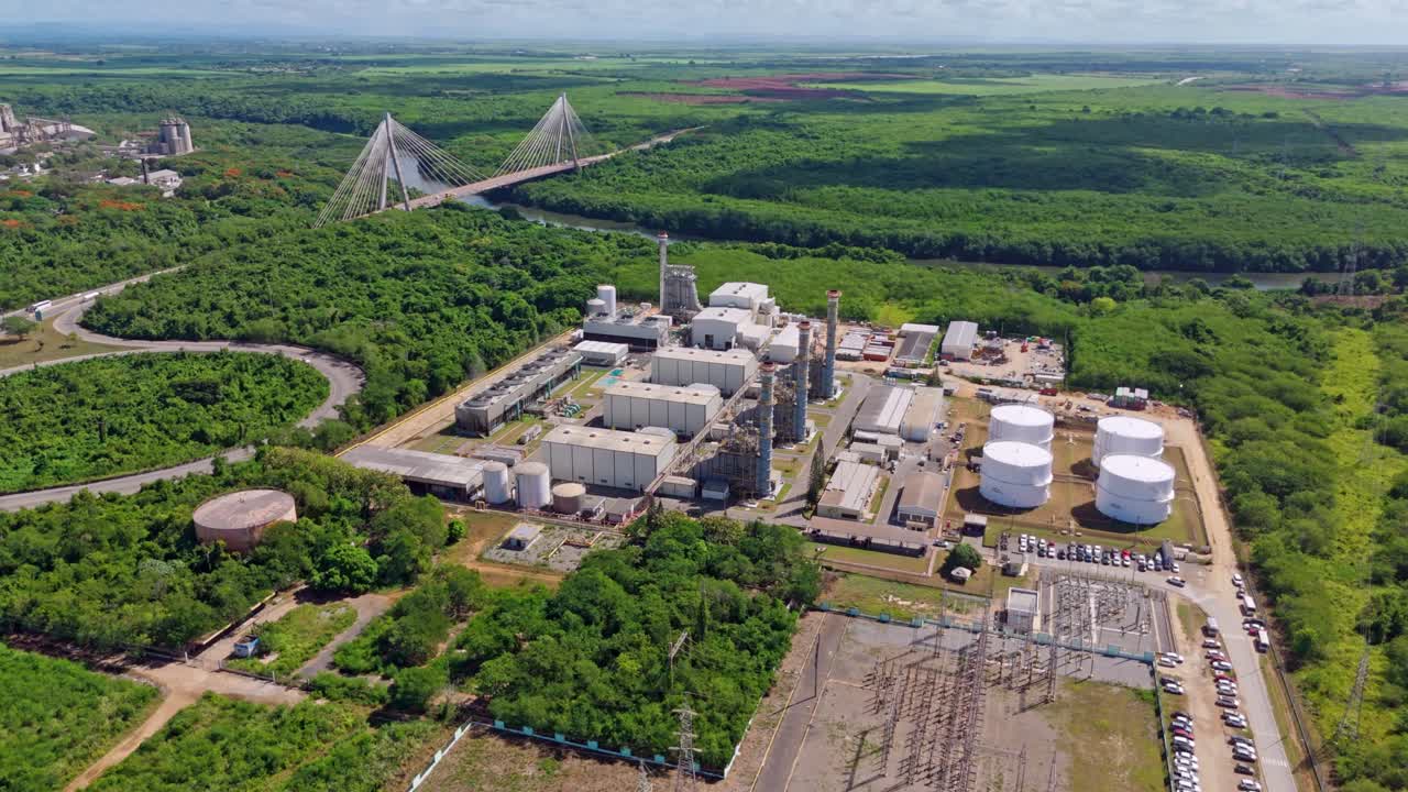 Energa 4 power plant, storage tanks, and substation, Mauricio Baez Bridge and cement plant visible in background, surrounded by lush forest, Dominican Republic. Aerial drone