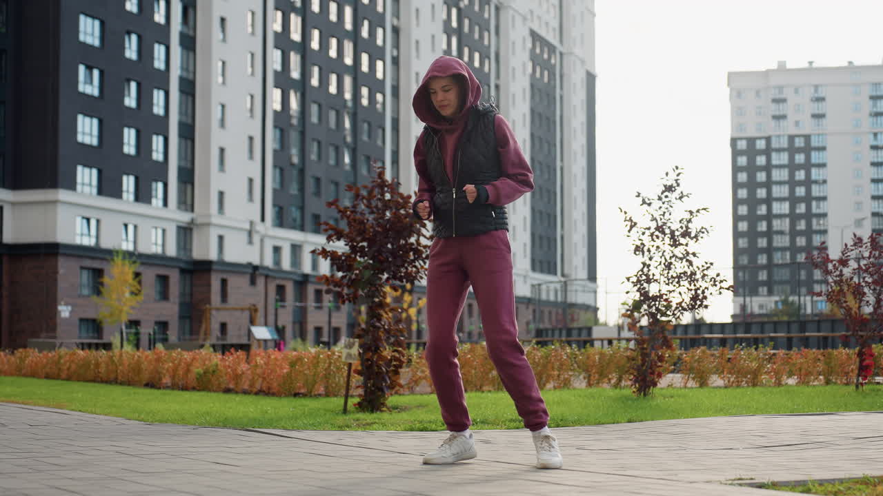 Female jogger in sports wear jumping during warm up exercise with excitement on face outdoor under bright sun amid flowers and skyscrapers on paved plaza lined with autumn shrubs and city skyline
