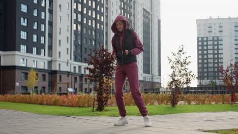 female jogger in sports wear jumping during warm up exercise with excitement on face outdoor under bright sun amid flowers and skyscrapers on paved plaza lined with autumn shrubs and city skyline
