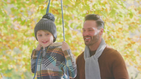 father pushing son having fun on rope swing in autumn garden