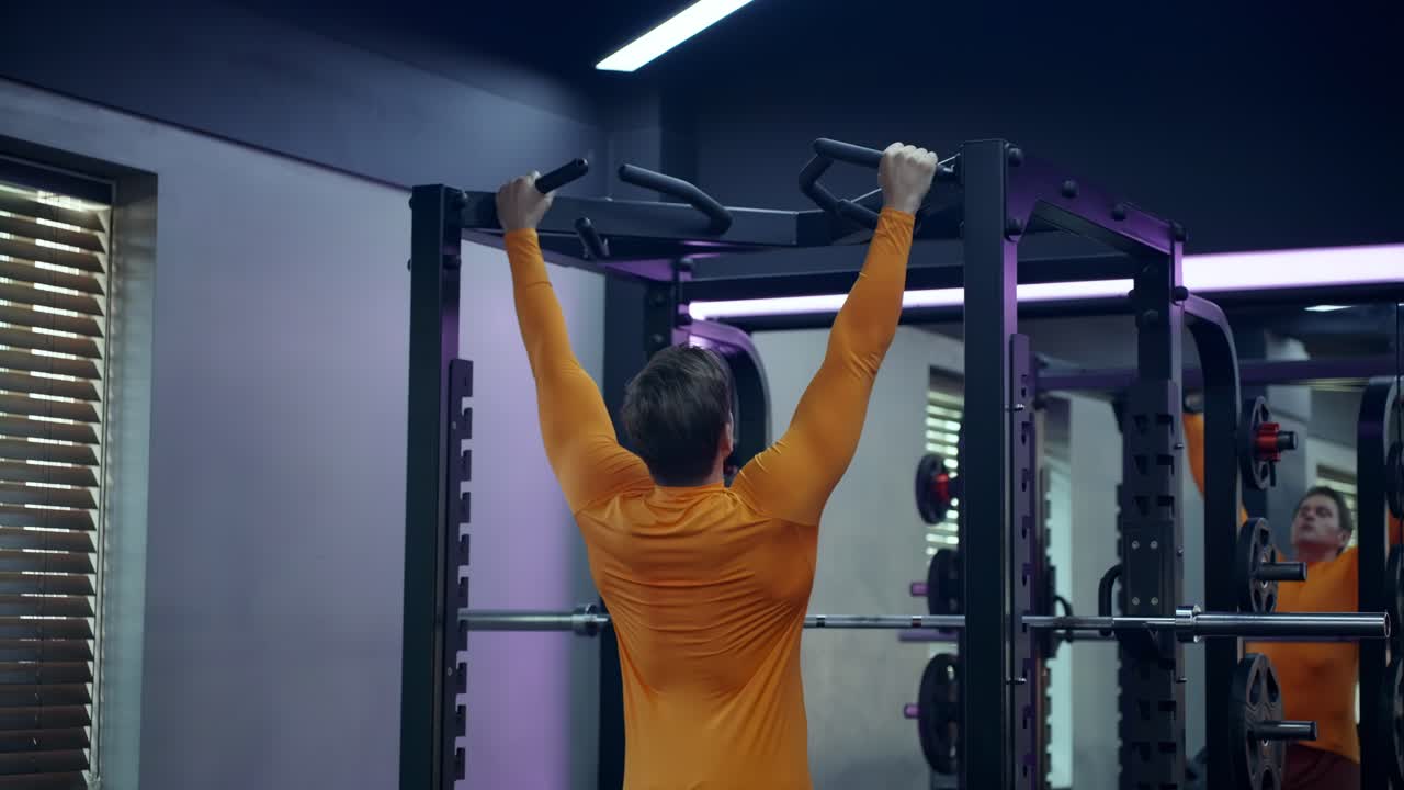 Crossfit athlete doing pull-ups at the gym - rear view. Handsome man in orange sportwear doing functional training on horizontal bar - slow motion