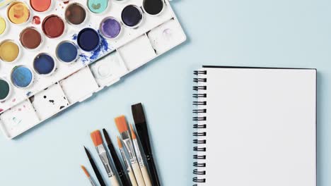 overhead view of notebook, brushes and paints on blue background, in slow motion