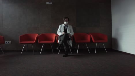 Young-doctor-sitting-in-a-red-hospital-chair-wearing-a-white-coat-and-respirator-working-with-documents