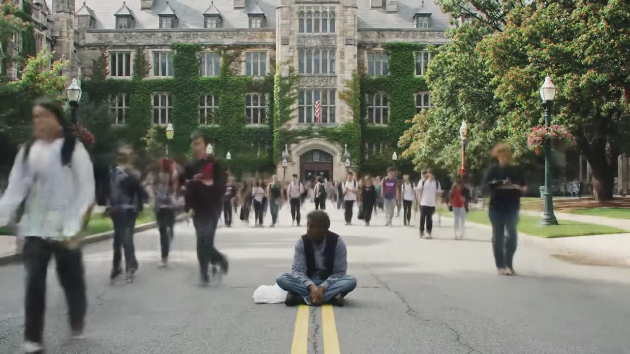 In a bustling university campus, a student sits calmly on the street as fellow students hurry by, highlighting the contrast between individual moments and the busy college life around him.