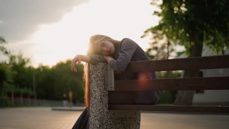 lady seated at edge of bench, resting with head leaning on arm, as the sun reflects soft light around her, someone walks in the background with a serene outdoor park environment surrounding her