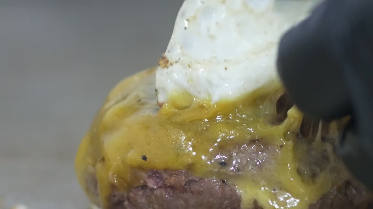 Primo piano delle mani guantate di uno chef mentre regola la posizione di un uovo fritto sopra un hamburger gourmet in cottura