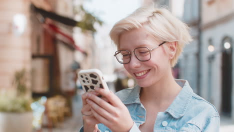 smiling blond woman using smartphone typing texting scrolling social media app on city town street