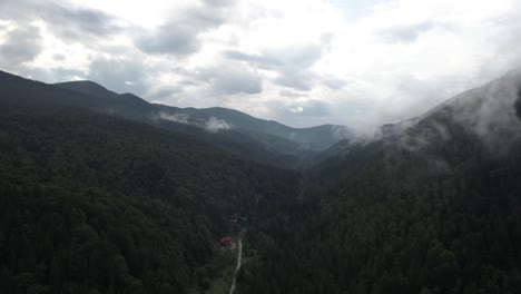 Flying-into-valley-covered-in-pine-forest-and-low-hanging-clouds-with-lone-house-standing-at-the-end-of-a-road