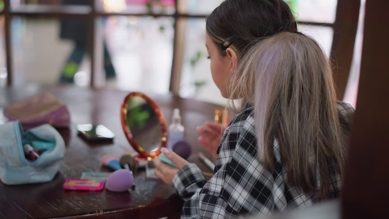 vista trasera de una dama aplicando maquillaje en polvo en la cara mientras se mira en el espejo, aseo personal, rutina de belleza capturada con herramientas de maquillaje esparcidas en la mesa