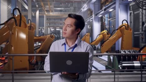 asian male professional worker standing with his laptop with industrial robotic arms working, he observes by looking around before he come to concentrating with his laptop