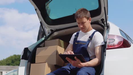 the courier sits on the edge of the open trunk, filled with boxes of goods and fills out documents. delivery of goods home