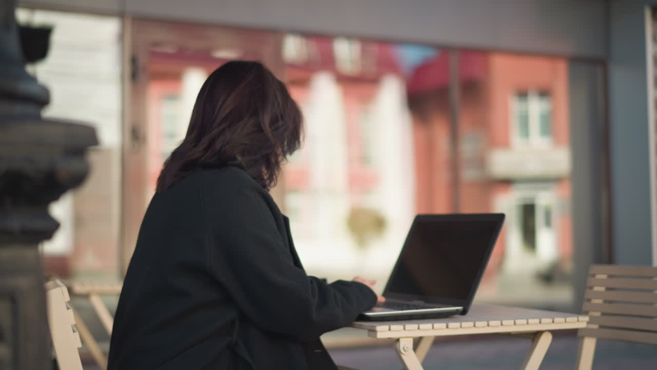 Elegant lady working on laptop outdoors, wearing stylish black coat, seated in urban environment with decorative black pillar and modern glass building reflecting red architecture in the background