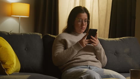 Young Woman Spending Evening At Home Sitting On Sofa With Mobile Phone Scrolling Through Internet Or Social Media 2