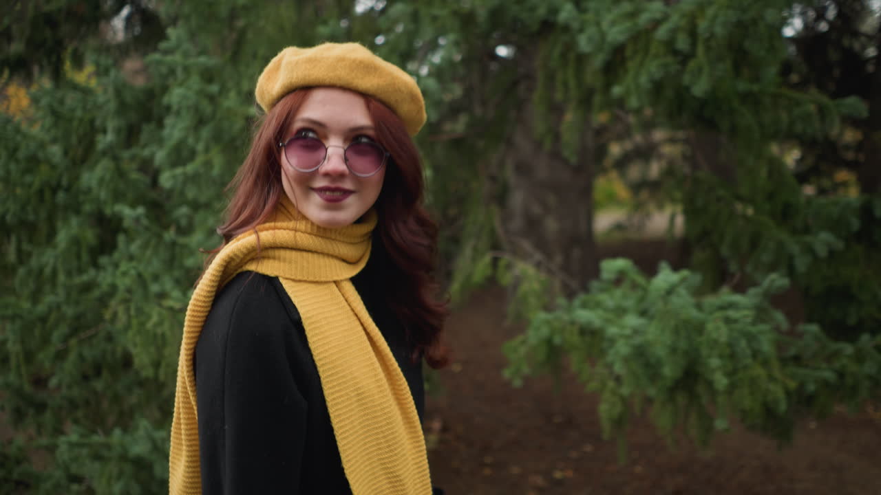 Stilfull kvinne med langt rødt hår og malte lepper, iført gul beret og skjerf, ser glad ut mens hun betrakter noe i bakgrunnen, med solbriller som bidrar til hennes elegante utseende i en frodig park.